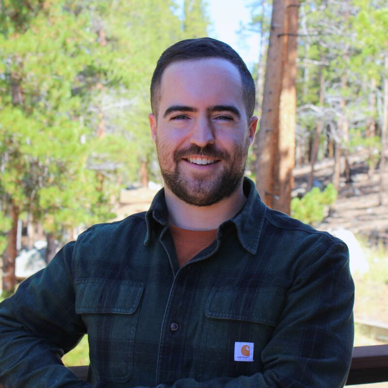 The image shows a man with a beard and short brown hair, wearing a green and black plaid shirt over an orange t-shirt. He is smiling and looking directly at the camera. The background features a forest with tall trees and a blurred, sunny sky. The man appears to be outdoors, possibly on a deck or porch, given the railing visible in the lower portion of the frame.